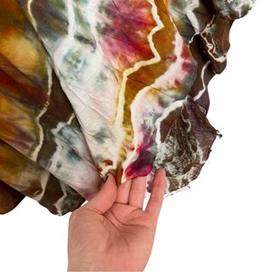 May include: A close-up of a tie-dye fabric with brown, orange, pink, and white colors. The fabric is folded and has a frayed edge.