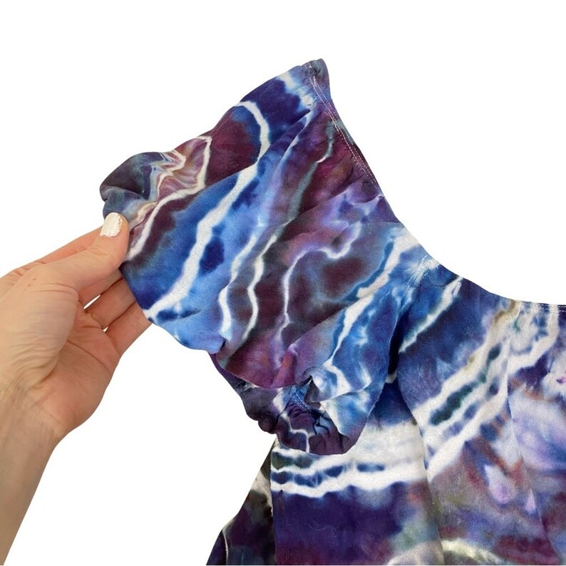 May include: A tie-dye off-the-shoulder top with a ruffled neckline in shades of blue, purple, and white.