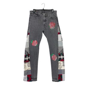 May include: Gray denim jeans with patchwork detailing on the sides and three embroidered red roses. The patchwork includes various patterns in red, white, black, and gray. The jeans are hanging on a black hanger.