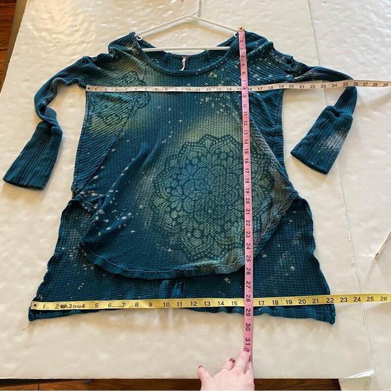 May include: Teal blue waffle knit long-sleeved shirt with a white mandala design. The shirt has a distressed look with white splatter paint accents.