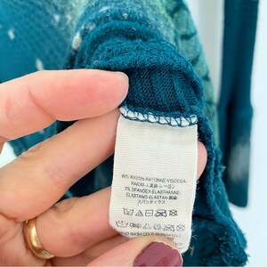 May include: Teal blue long-sleeved shirt with a white and teal tie-dye pattern. The shirt has a tag with the following information: 95% Rayon, 3% Spandex, Hand Wash Cold Separately.