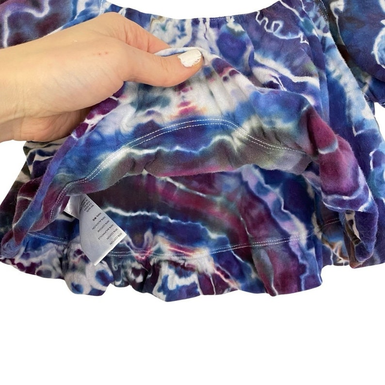 May include: A purple, blue, and white tie-dye off-the-shoulder top with a ruffled hem. The top is made of a soft, stretchy fabric and has a relaxed fit.