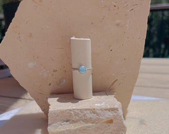 May include: A silver ring with a small, round, light blue gemstone. The ring is displayed on a white cylindrical object, set against a backdrop of beige stone blocks and greenery. The ring's simple design highlights the gemstone.