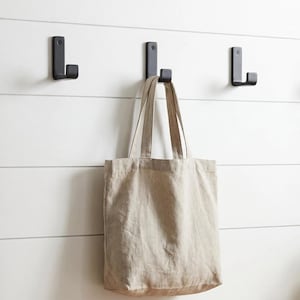 May include: Four black metal L-shaped hooks on a white slatted wall. A beige canvas tote bag hangs from one hook. A wooden bench and a potted plant with round green leaves are in the foreground.