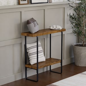 May include: A two-tiered wooden side table with black metal legs. The top shelf holds a gray pillow with fringe and a small white pillow with black stripes. A larger white pillow with black dotted lines rests on the bottom shelf.