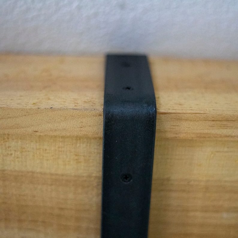 May include: Close-up of a black metal bracket attached to a light brown wooden shelf.