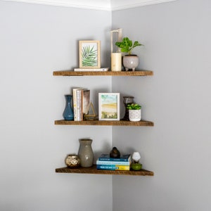 Könnte beinhalten: Drei Holzregale, die in einer Ecke montiert sind, mit verschiedenen Gegenständen darauf, darunter Bücher, Pflanzen, Vasen und gerahmte Kunstwerke. Die Regale sind aus recyceltem Holz und haben einen rustikalen Look.