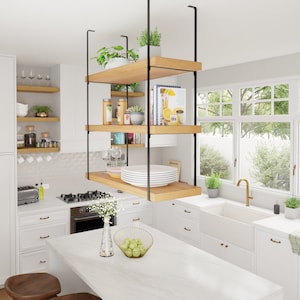 May include: A white kitchen with a floating shelf unit made of wood and black metal. The shelf unit is filled with various items, including plants, books, and dishes. The kitchen also features a white farmhouse sink, a white worktop, and white cabinets.