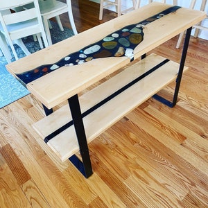 May include: A wooden console table with a black metal frame and a clear epoxy river table top with embedded stones. The table has a lower shelf.