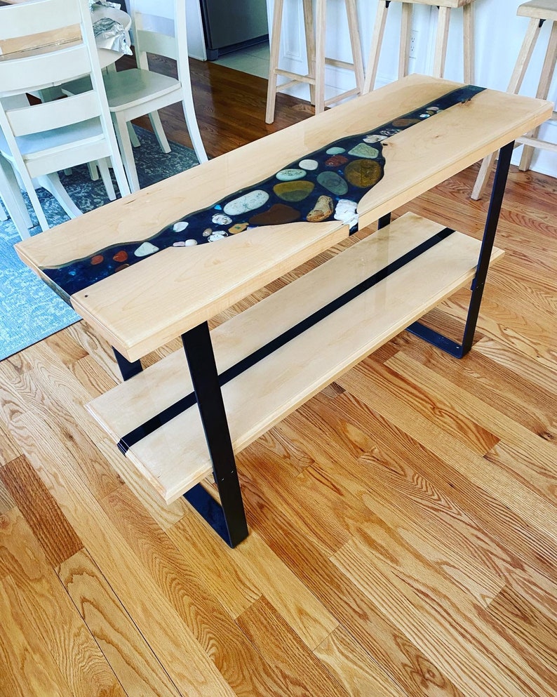May include: A wooden console table with a black metal frame and a clear epoxy river table top with embedded stones. The table has a lower shelf.
