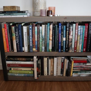 May include: A wooden bookshelf with two shelves, filled with books about art and artists. The books are organised by author and subject. The bookshelf is made of dark wood with black metal accents.