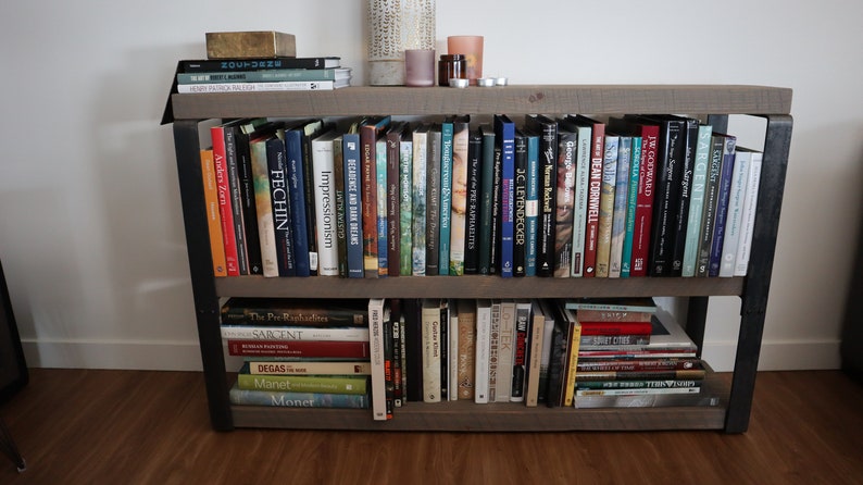 May include: A wooden bookshelf with two shelves, filled with books about art and artists. The books are organised by author and subject. The bookshelf is made of dark wood with black metal accents.