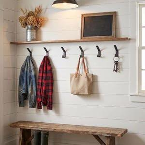 May include: A country-style hallway with a wooden bench, a shelf, and black hooks. A denim jacket, a red plaid shirt, and a canvas tote bag hang from the hooks. A framed chalkboard and a metal vase with dried flowers are on the shelf.