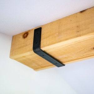 May include: A close-up of a wooden beam with a black metal bracket attached to it. The beam is light brown and the bracket is made of metal.
