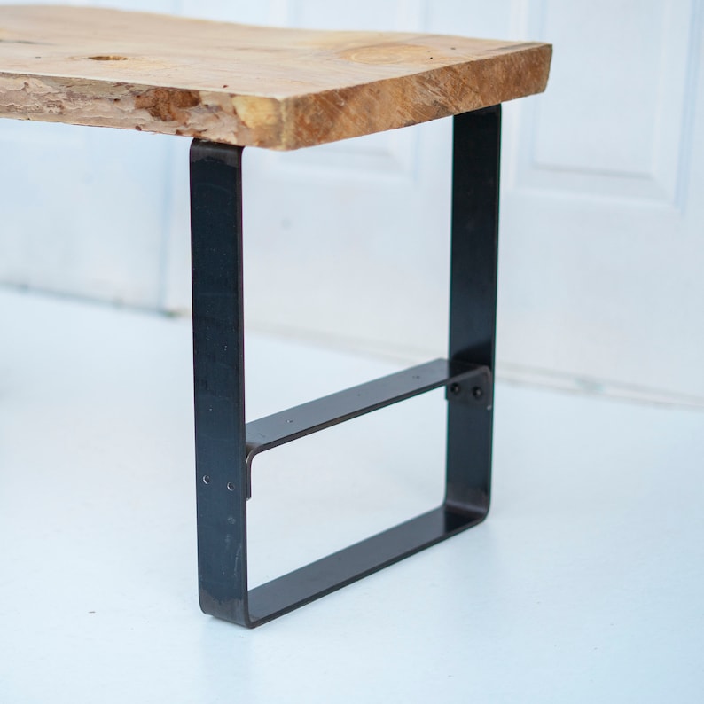 May include: A wooden table top with a live edge sits on black metal legs. The legs are rectangular and have a simple design.