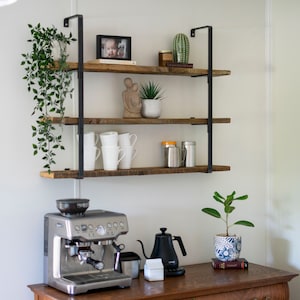 Puede incluir: Una estación de café en casa con una máquina de espresso de acero inoxidable, una tetera negra y una pequeña planta en una maceta con estampado azul y blanco. Estantes de madera con soportes de metal negro sostienen tazas, una foto enmarcada y objetos decorativos.