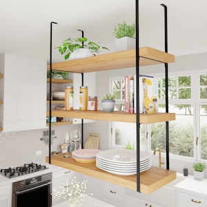 May include: A kitchen featuring a floating shelf unit crafted from wood and black metal. The shelf unit comprises two shelves and is suspended from the ceiling. The shelves are filled with various items, including plants, books, and dishes. The kitchen also boasts a white countertop, a stainless steel stove, and a window with a view of trees.