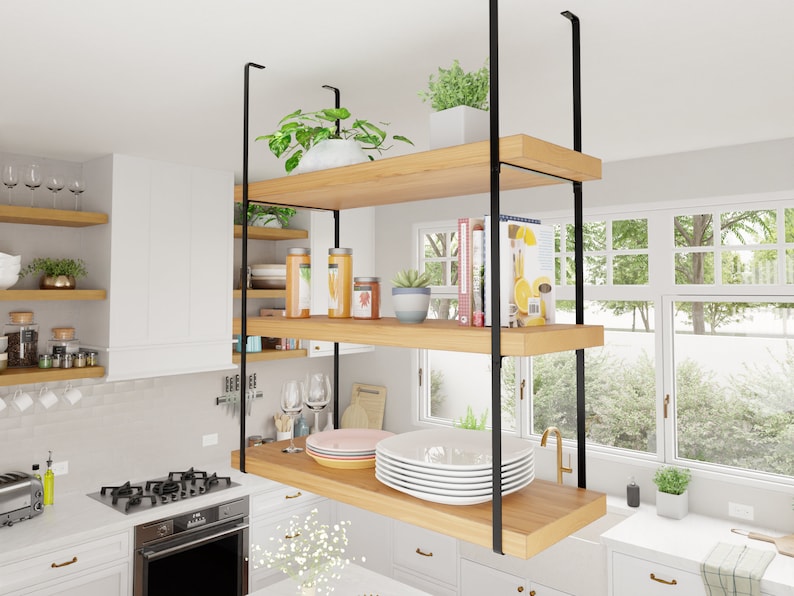 May include: A kitchen featuring a floating shelf unit crafted from wood and black metal. The shelf unit comprises two shelves and is suspended from the ceiling. The shelves are filled with various items, including plants, books, and dishes. The kitchen also boasts a white countertop, a stainless steel stove, and a window with a view of trees.