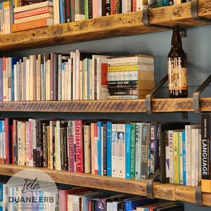 K&ouml;nnte beinhalten: Ein rustikales Holzregal mit drei Regalbrettern, die jeweils mit B&uuml;chern gef&uuml;llt sind. Die Regalbretter werden von schwarzen Metallkonsolen getragen. Eine Flasche Bier steht auf dem oberen Regal.