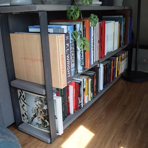 May include: A black metal bookshelf with two shelves filled with books. The bookshelf is in a corner of a room with a grey sofa and a wooden floor. The books are mostly hardback and have a variety of colours and titles.