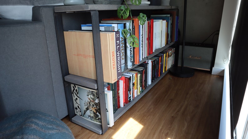 May include: A black metal bookshelf with two shelves filled with books. The bookshelf is in a corner of a room with a grey sofa and a wooden floor. The books are mostly hardback and have a variety of colours and titles.