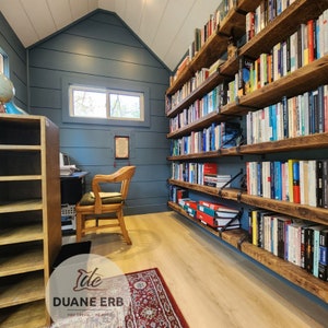 K&ouml;nnte beinhalten: Ein rustikales Holzregal mit mehreren Regalbrettern, die mit B&uuml;chern gef&uuml;llt sind. Das Regal ist an einer Wand in einem Raum mit blauen W&auml;nden und einem Holzschreibtisch mit Stuhl montiert.