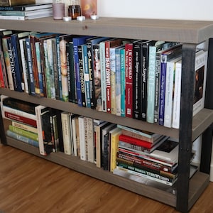 May include: A grey wooden bookshelf with black metal accents, filled with books. The bookshelf has two shelves and is standing on a wooden floor.