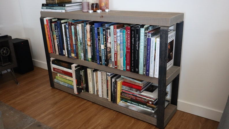May include: A grey wooden bookshelf with black metal accents, filled with books. The bookshelf has two shelves and is standing on a wooden floor.
