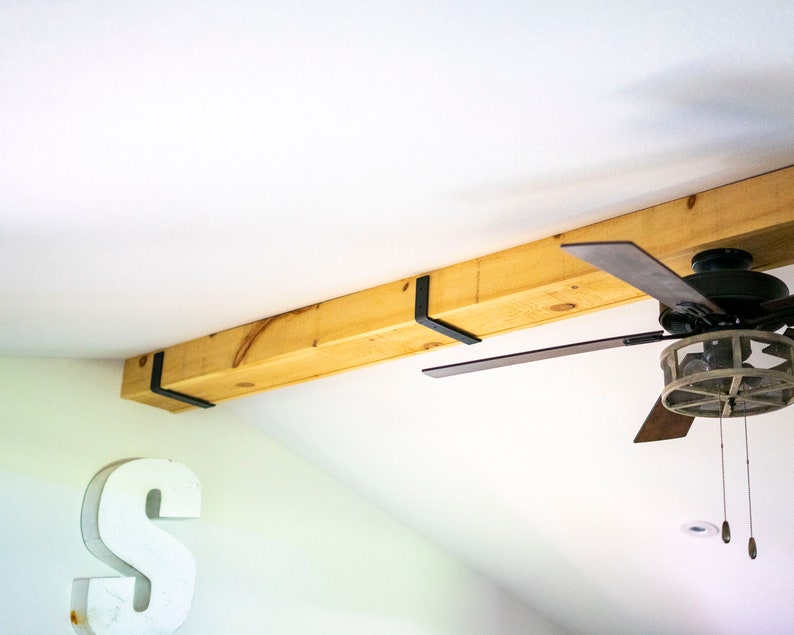 May include: A wooden beam with black metal brackets attached to the ceiling. A ceiling fan with brown blades is mounted to the ceiling.