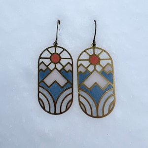 Op de afbeelding: Een paar gouden oorbellen met een geometrisch ontwerp met een gestileerde berg scène met een rode zon, blauwe bergen en witte sneeuw. De oorbellen zijn gemaakt van metaal en hebben een glanzende afwerking.