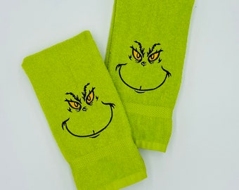 May include: Two lime green hand towels with embroidered Grinch faces. Each towel features a black outline of the Grinch's face with yellow eyes and a wide smile. The towels are made of a soft, absorbent material.
