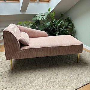Può includere: Una chaise longue in velluto rosa con gambe dorate si trova su un tappeto color crema in una stanza con un lucernario. La chaise longue ha un cuscino ed è inclinata verso destra.