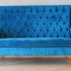 May include: A blue velvet tufted sofa with wooden legs. The sofa has a high back and wingback arms.
