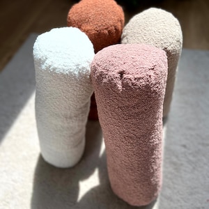May include: Four cylindrical pillows in different colors: white, light brown, pink, and beige. The pillows are made of a soft, textured fabric and are arranged in a circle.