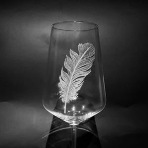 Könnte beinhalten: Ein klares Weinglas mit langem Stiel, das ein geätztes Federdesign aufweist. Die Feder ist mit feinen Linien detailliert, wodurch ein strukturiertes Aussehen entsteht. Das Glas steht vor einem dunklen Hintergrund, der die zarten Details der Feder hervorhebt.