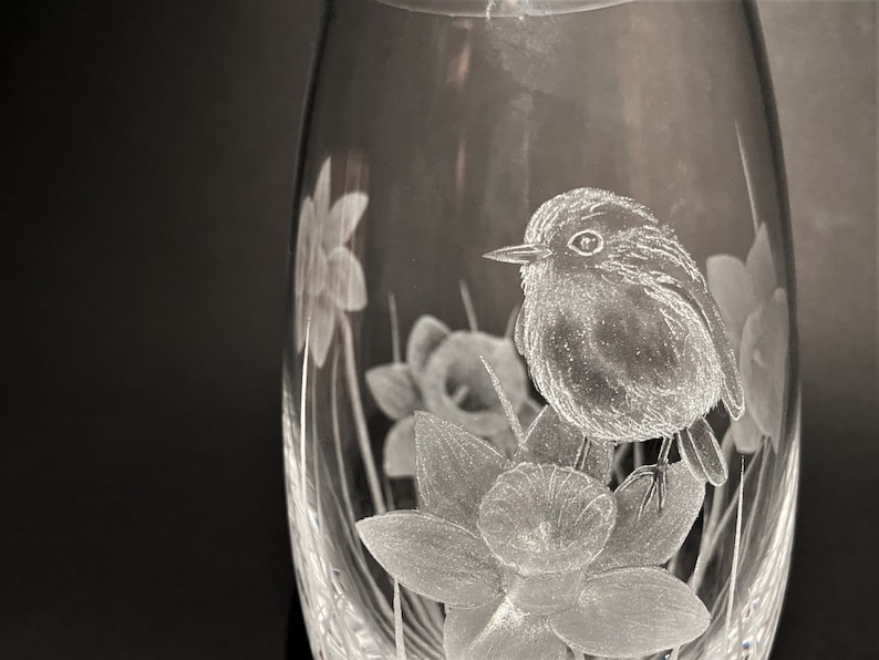 K&ouml;nnte beinhalten: Eine klare Glasvase mit einem ge&auml;tzten Design eines Rotkehlchens, das auf einer Narzisse sitzt. Die Vase ist oval geformt und hat ein zartes, filigranes Design.