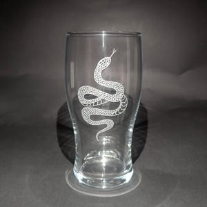 May include: Clear glass beer glass featuring a white etched snake design. The snake is coiled and detailed, with a forked tongue. The glass has a wide top and a narrow base, suitable for beer or other beverages.