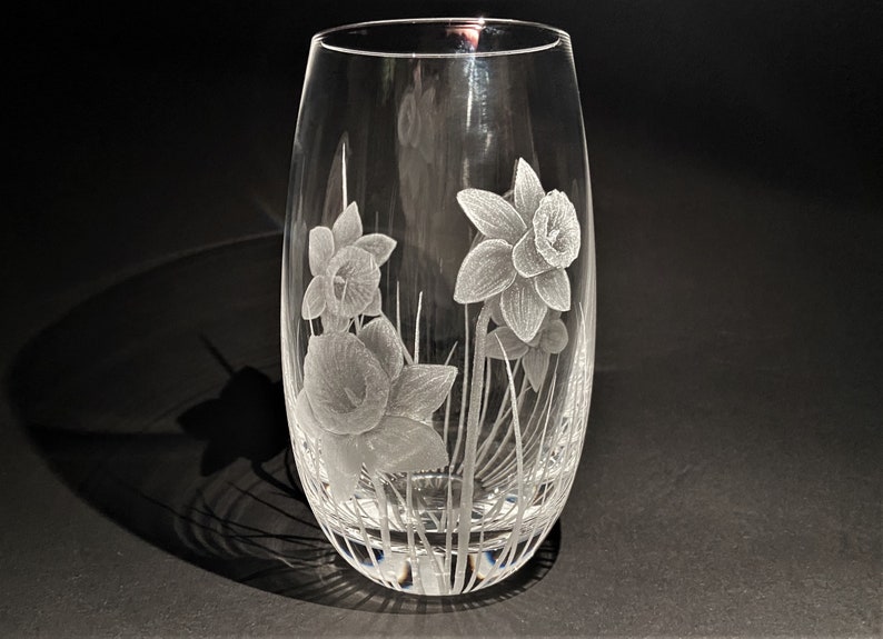 K&ouml;nnte beinhalten: Eine klare Glasvase mit einem ge&auml;tzten Design von Narzissen. Die Vase hat einen runden Boden und eine hohe, schlanke Form.