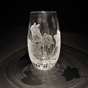 K&ouml;nnte beinhalten: Eine klare Glasvase mit einem gravierten Design eines Vogels und Narzissen. Die Vase hat einen runden Boden und einen hohen, schlanken K&ouml;rper.