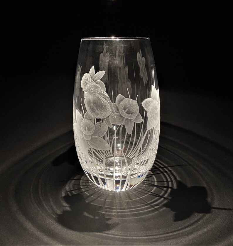 K&ouml;nnte beinhalten: Eine klare Glasvase mit einem gravierten Design eines Vogels und Narzissen. Die Vase hat einen runden Boden und einen hohen, schlanken K&ouml;rper.