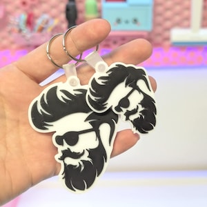 Puede incluir: Dos llaveros con la silueta en blanco y negro de un hombre barbudo con gafas de sol. Los llaveros están unidos por un anillo de metal plateado y una correa de plástico transparente. El fondo es rosa borroso.