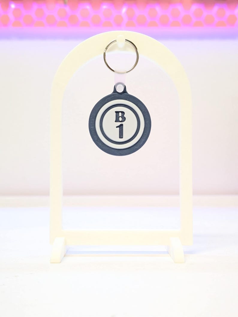 May include: A cream-colored arched display stand holding a navy blue and white keychain. The keychain has the letter "B" and the number "1" printed on it. The stand has a rectangular base.