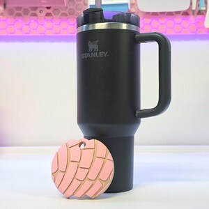 Puede incluir: Un vaso t&eacute;rmico negro Stanley con asa y un llavero rosa en forma de concha. El llavero tiene un contorno marr&oacute;n claro y un agujero para sujetarlo. El vaso tiene un borde plateado y una pajita negra.