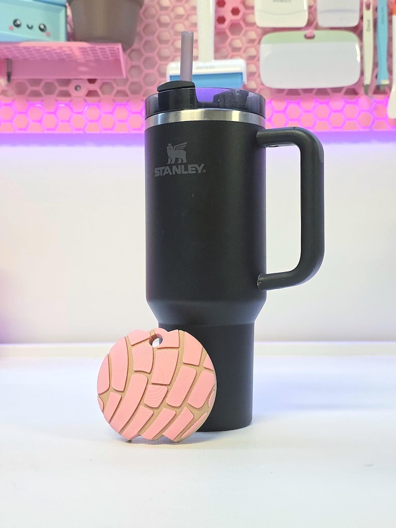 Puede incluir: Un vaso t&eacute;rmico negro Stanley con asa y un llavero rosa en forma de concha. El llavero tiene un contorno marr&oacute;n claro y un agujero para sujetarlo. El vaso tiene un borde plateado y una pajita negra.
