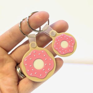 May include: Two donut-shaped keychains. Each keychain features a pink frosted donut with white sprinkles and a cream-colored center. The keychains are attached to silver-colored key rings.