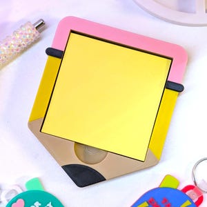May include: A yellow notepad in a pencil-shaped holder with a pink top and yellow sides. The holder has a tan and black pencil tip design. A pen and two keychains are also visible.