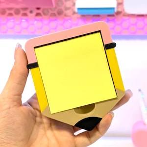 May include: A yellow sticky note pad shaped like a pencil, with a pink top and a tan bottom. The pad is held in a hand, and the background is a blurred display of other stationery items.