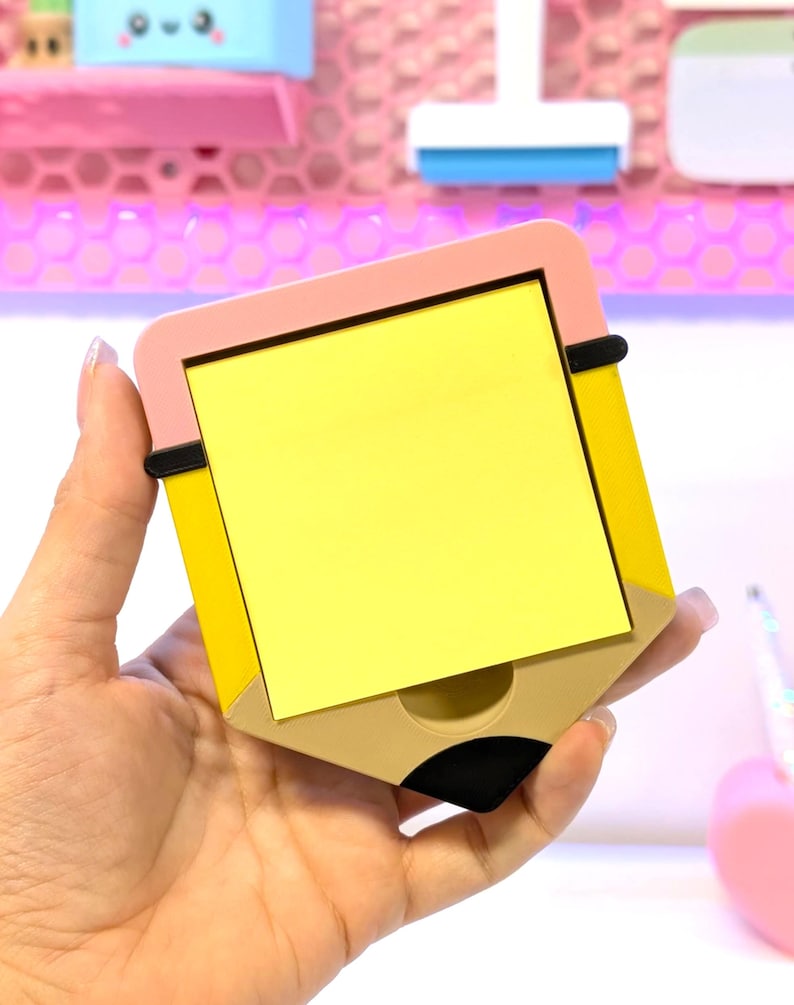 May include: A yellow sticky note pad shaped like a pencil, with a pink top and a tan bottom. The pad is held in a hand, and the background is a blurred display of other stationery items.