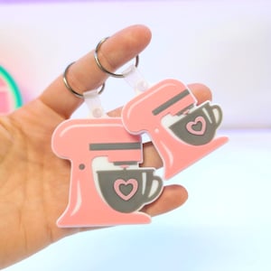 Op de afbeelding: Twee roze en grijze keukenmixer sleutelhangers met hart details. De sleutelhangers zijn bevestigd aan zilveren sleutelringen. Het mixerontwerp bevat een grijze kom met een hart en grijze accenten. De sleutelhangers worden vastgehouden door de vingers van een persoon.