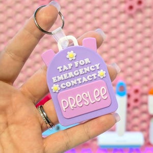 May include: A purple backpack-shaped keychain with the text "TAP FOR EMERGENCY CONTACT" and the name "Preslee" in white. The keychain has a silver keyring and is decorated with small yellow flowers.